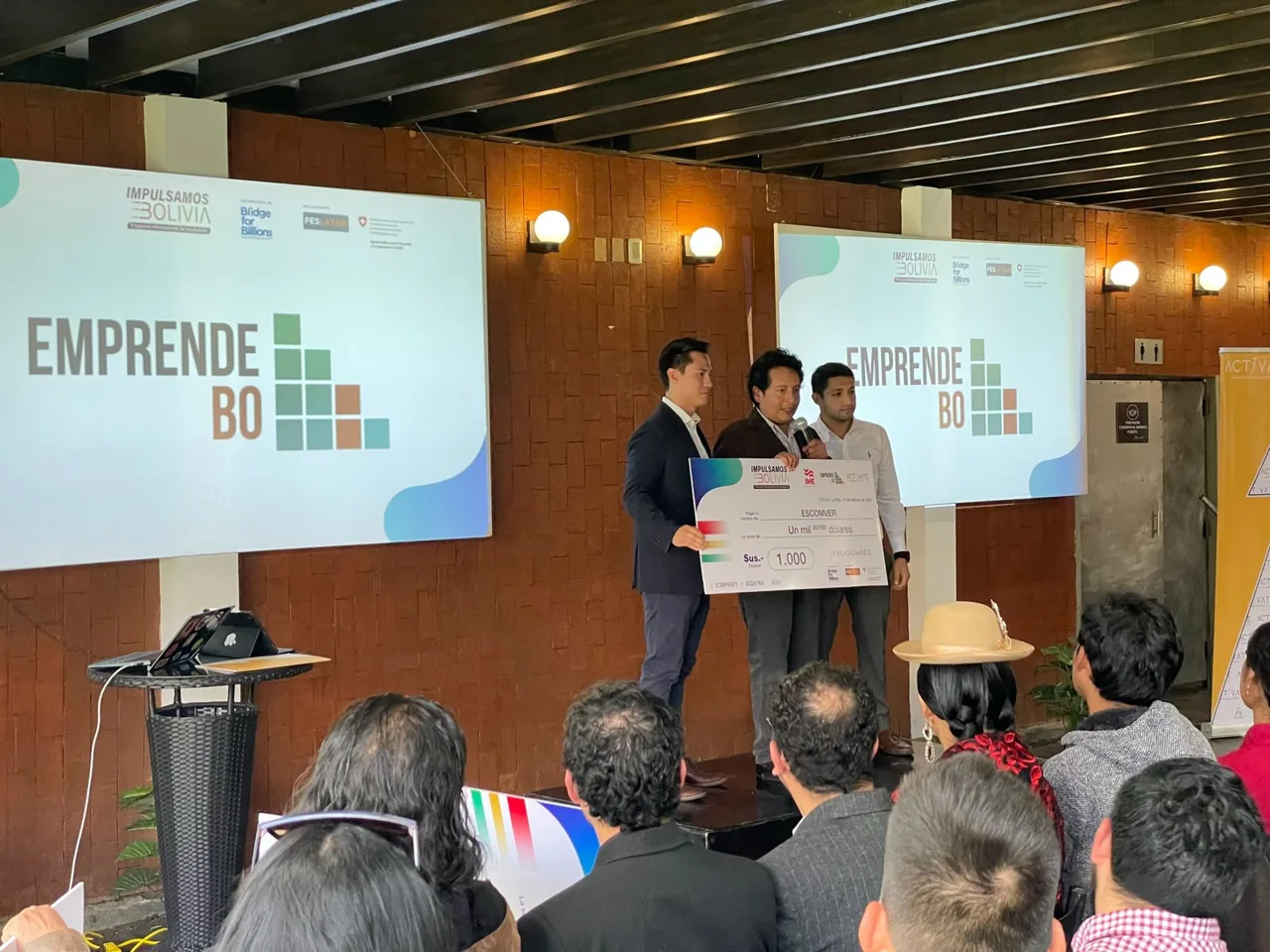 Entrega de reconocimiento en el evento de triple impacto, con EmprendeBO proyectado en las pantallas.